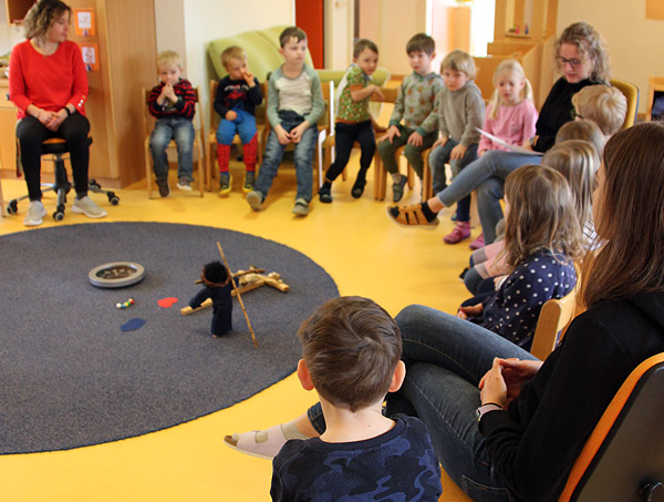 Die Kinder und Erzieher sitzen im Stuhlkreis. Die Figur in der Mitte des Stuhlkreuzes hat eine gebeugte Haltung, das Gesicht ist nicht zu erkennen. 